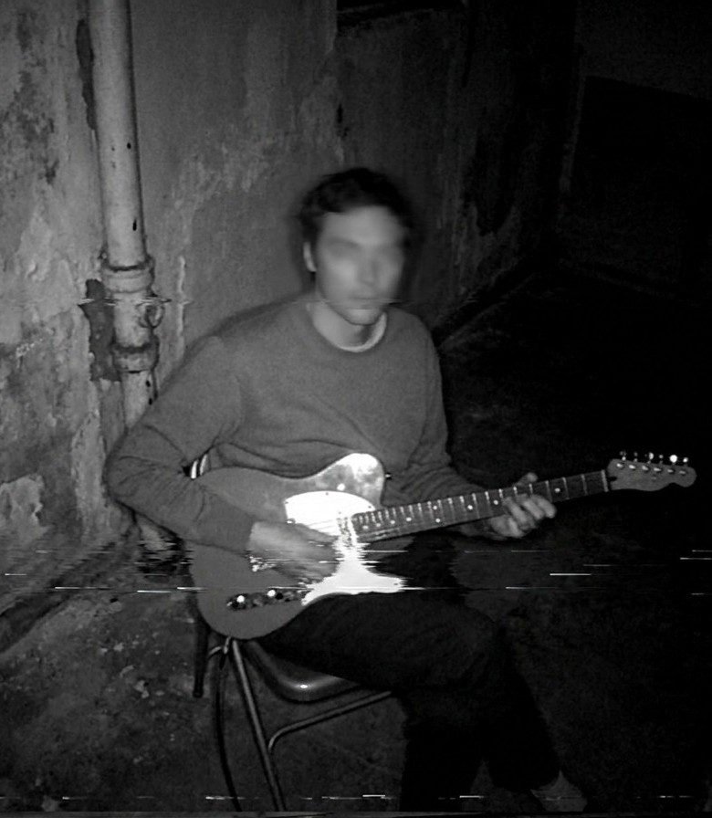 Robbie Burton, blurred, playing an electric guitar on a chair in a dim basement with peeling walls
