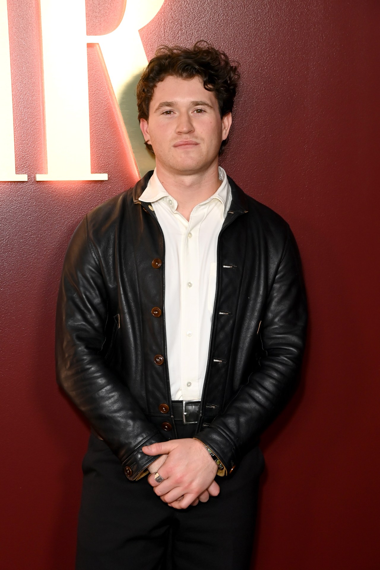 Rohan Campbell with curly hair, wearing a black leather jacket over a white shirt, standing against a dark red wall.