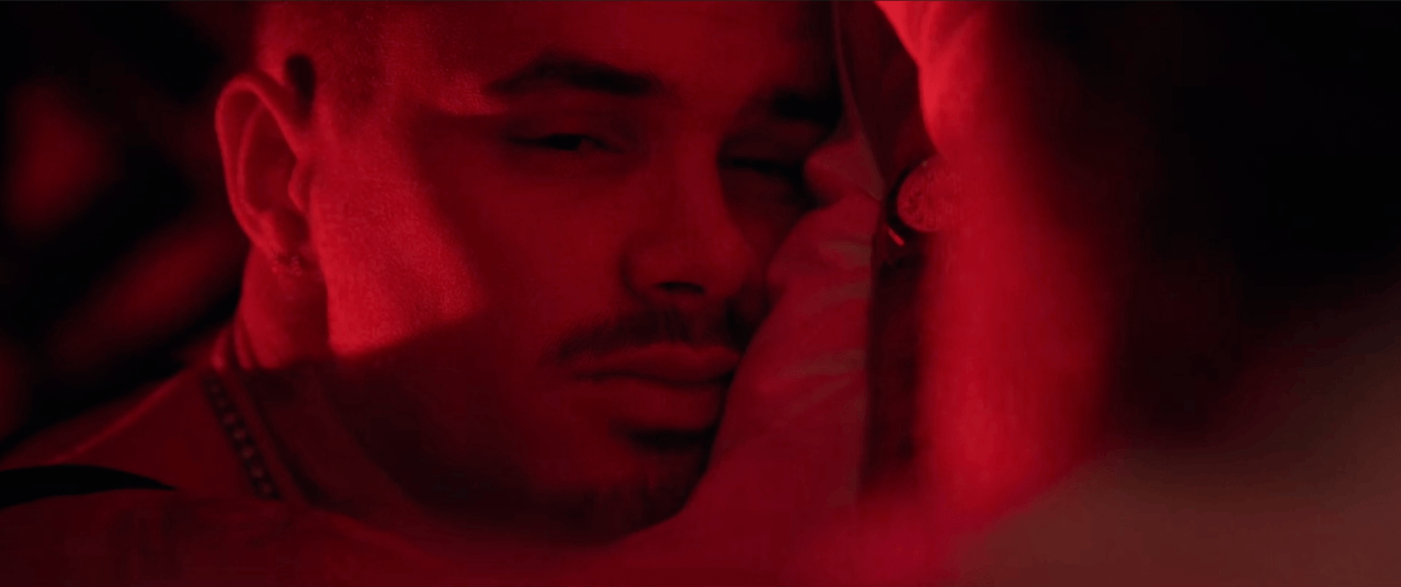 Young man's face lit in deep red, eyes closed, with a hand touching his cheek and a chain necklace visible at his throat.
