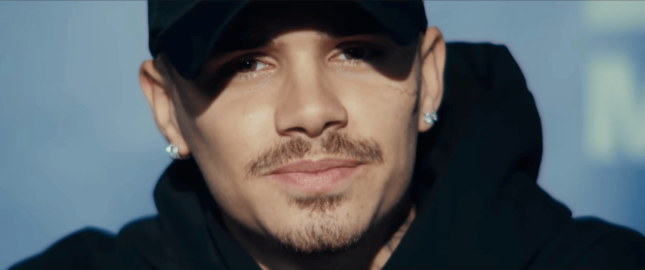 Young man in a black hoodie and black baseball cap looking slightly off-camera, stud earrings visible, against a soft blue background.