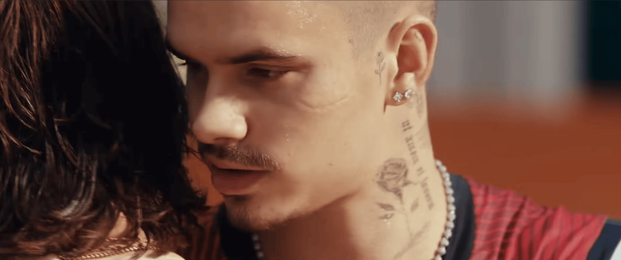 Close-up of a man with neck tattoos including a rose design leaning toward a dark-haired woman, both in profile.