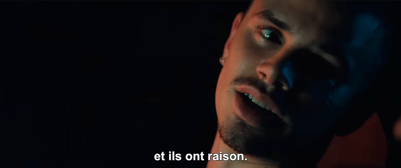 Man's face partially lit in blue against a black background, mouth open, with French subtitle "et ils ont raison" at bottom of frame.