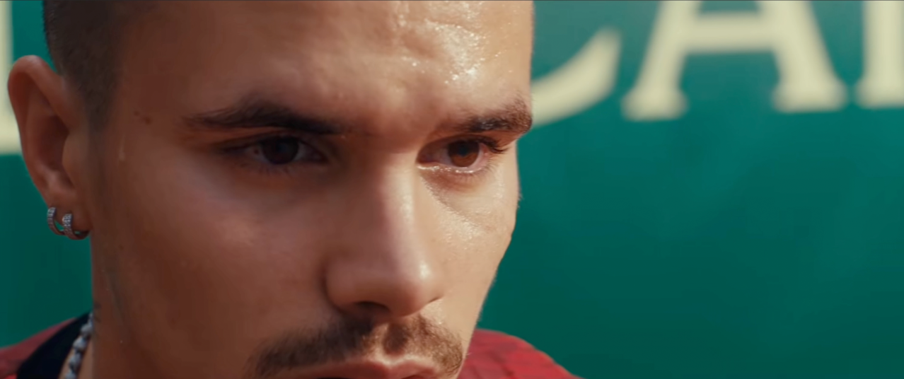 Extreme close-up of a young man's face, sweating, with a focused and intense expression, wearing a small hoop earring and a chain necklace.