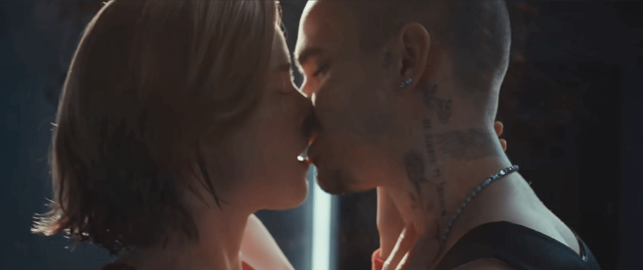 Two people leaning in to kiss in close-up; one has neck tattoos and a chain necklace, the other has a lip ring.