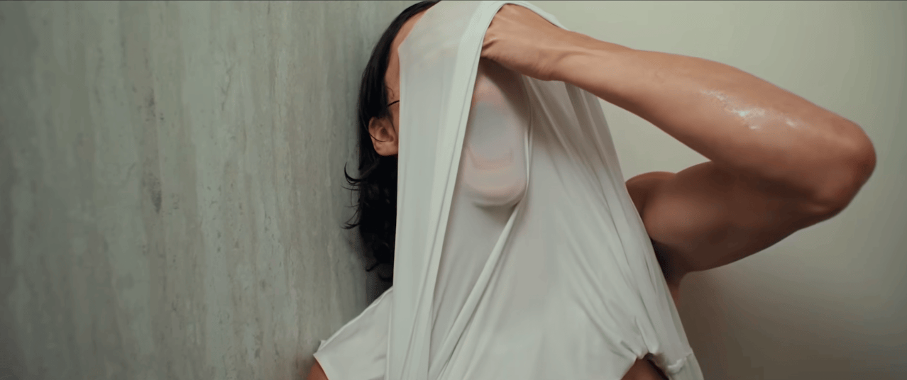 Woman with dark hair pulling a sheer white fabric over her face while leaning against a concrete wall, arm raised.