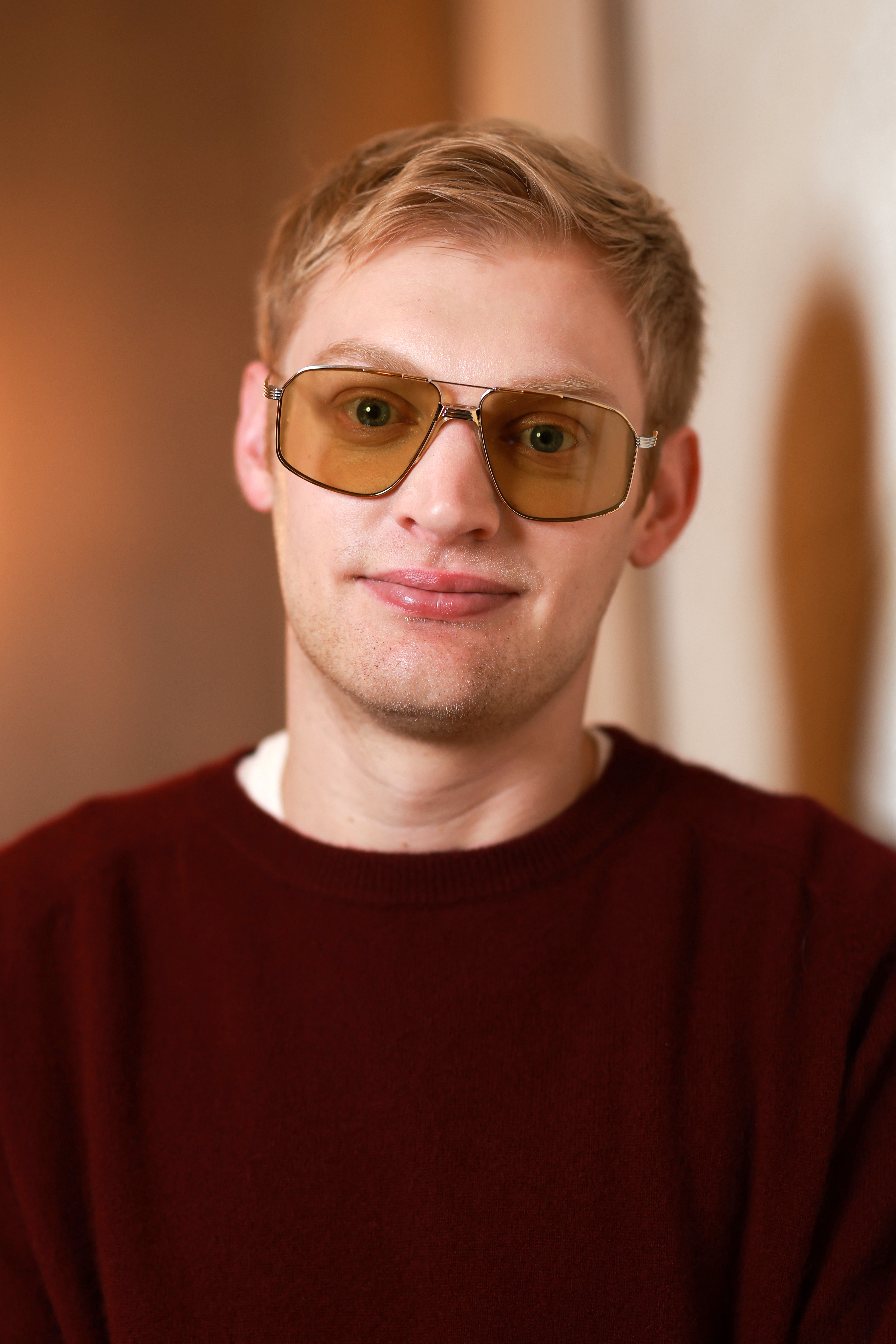 Young blond man wearing amber-tinted aviator sunglasses and a dark red crewneck sweater, smiling slightly at the camera.