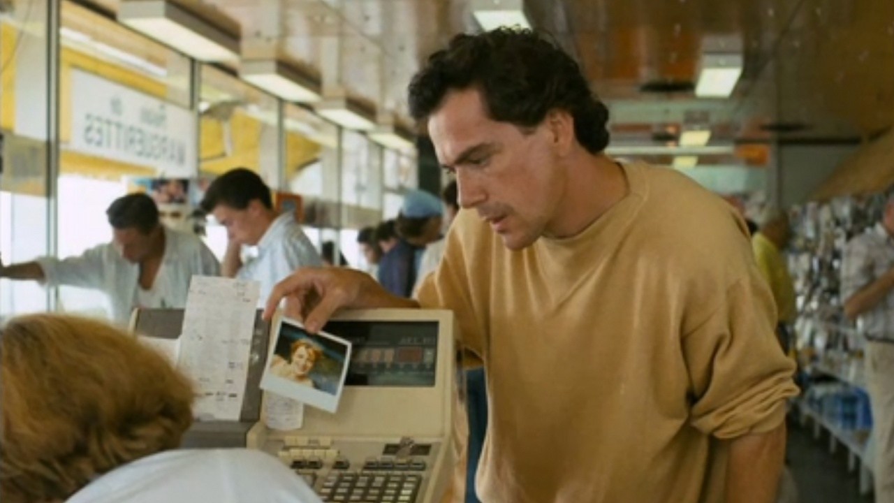 A dark-haired man in a yellow sweatshirt leans over a store checkout counter, holding a photograph out toward a cashier, with other shoppers visible in the background.