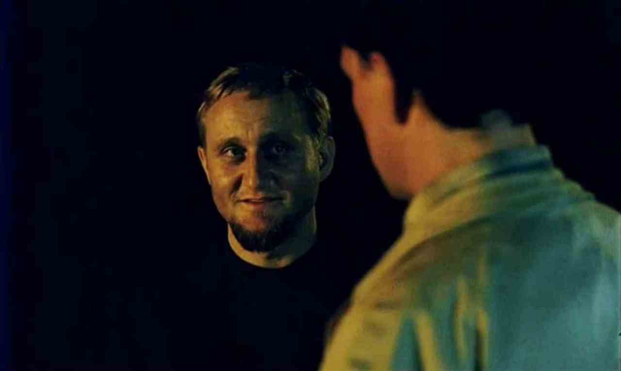 A blond man with a goatee smiles ominously in low, eerie light while facing another man seen from behind in the foreground, set against a near-black background.