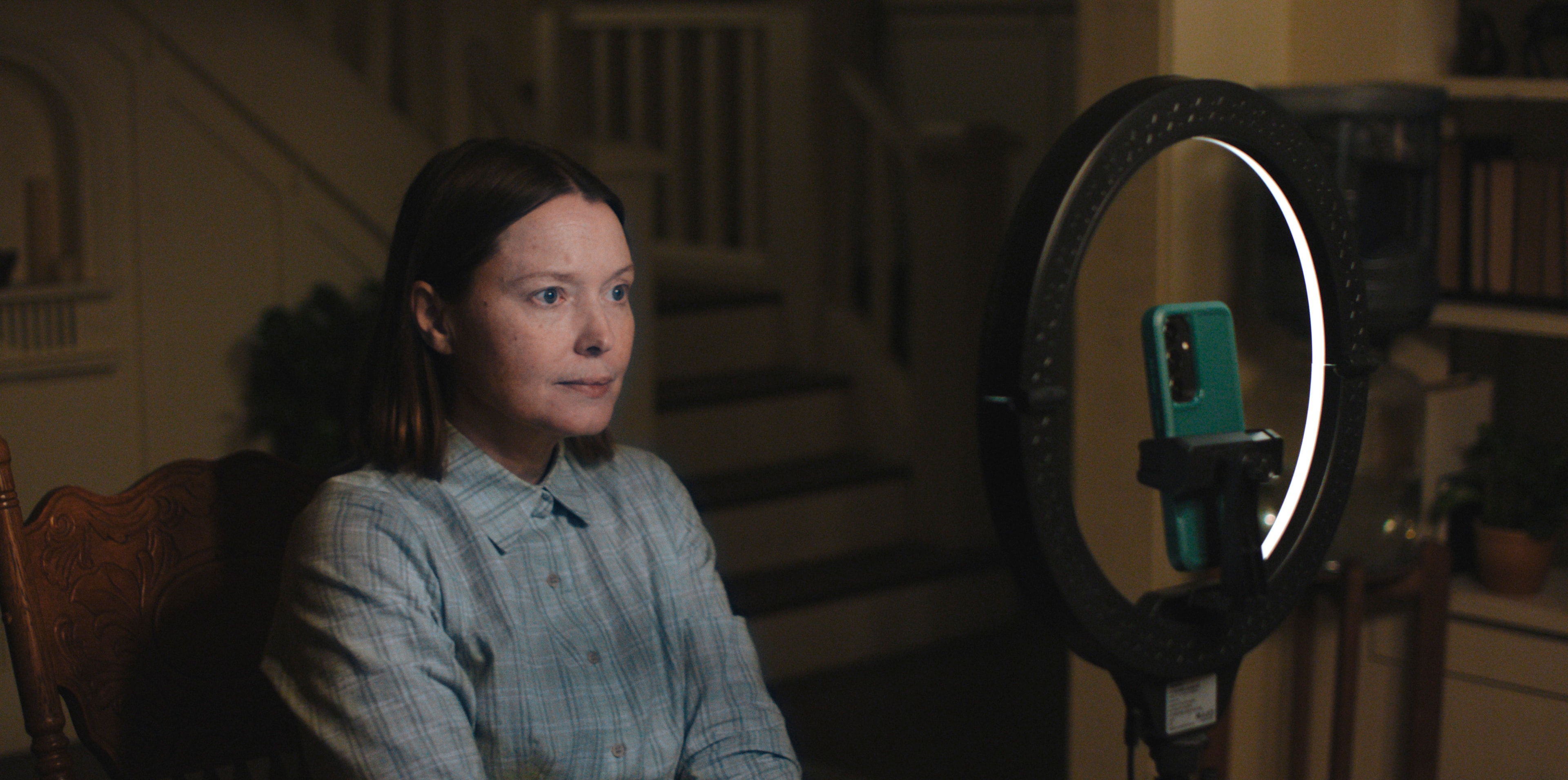 A woman with dark hair in a blue plaid shirt sits in a wooden chair, staring at a smartphone mounted on a ring light in a dimly lit house.