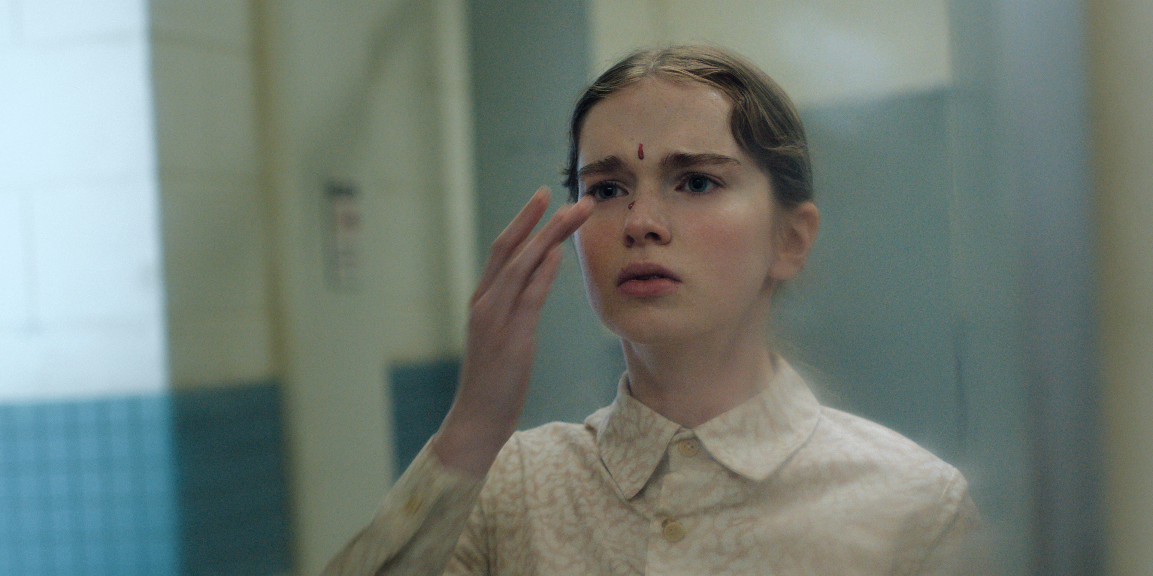 A teenage girl in a pale patterned blouse raises her hand toward her face, a small drop of blood visible on her forehead, looking startled.