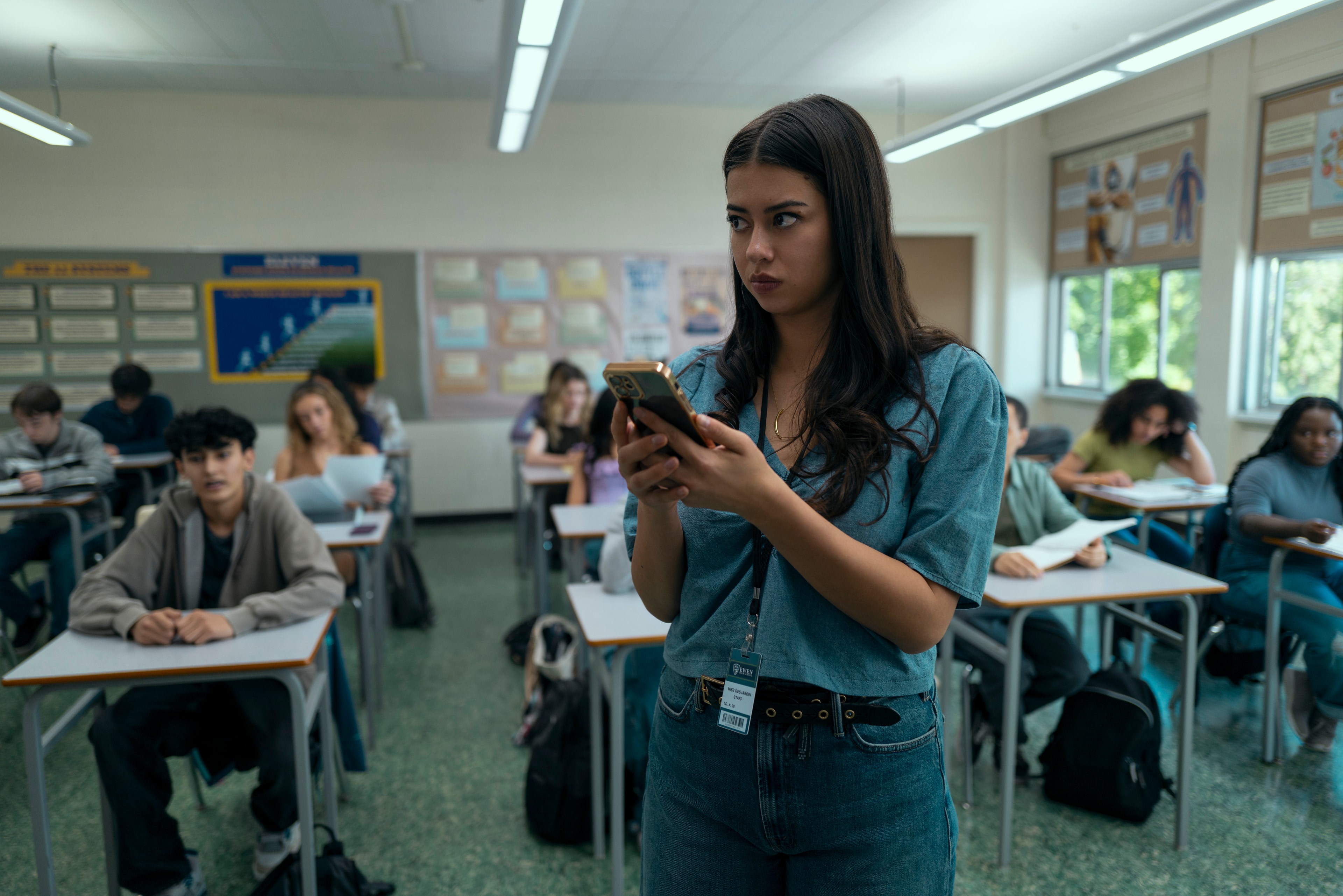 A young woman with long dark hair holds a smartphone at the front of a high school classroom full of seated students.