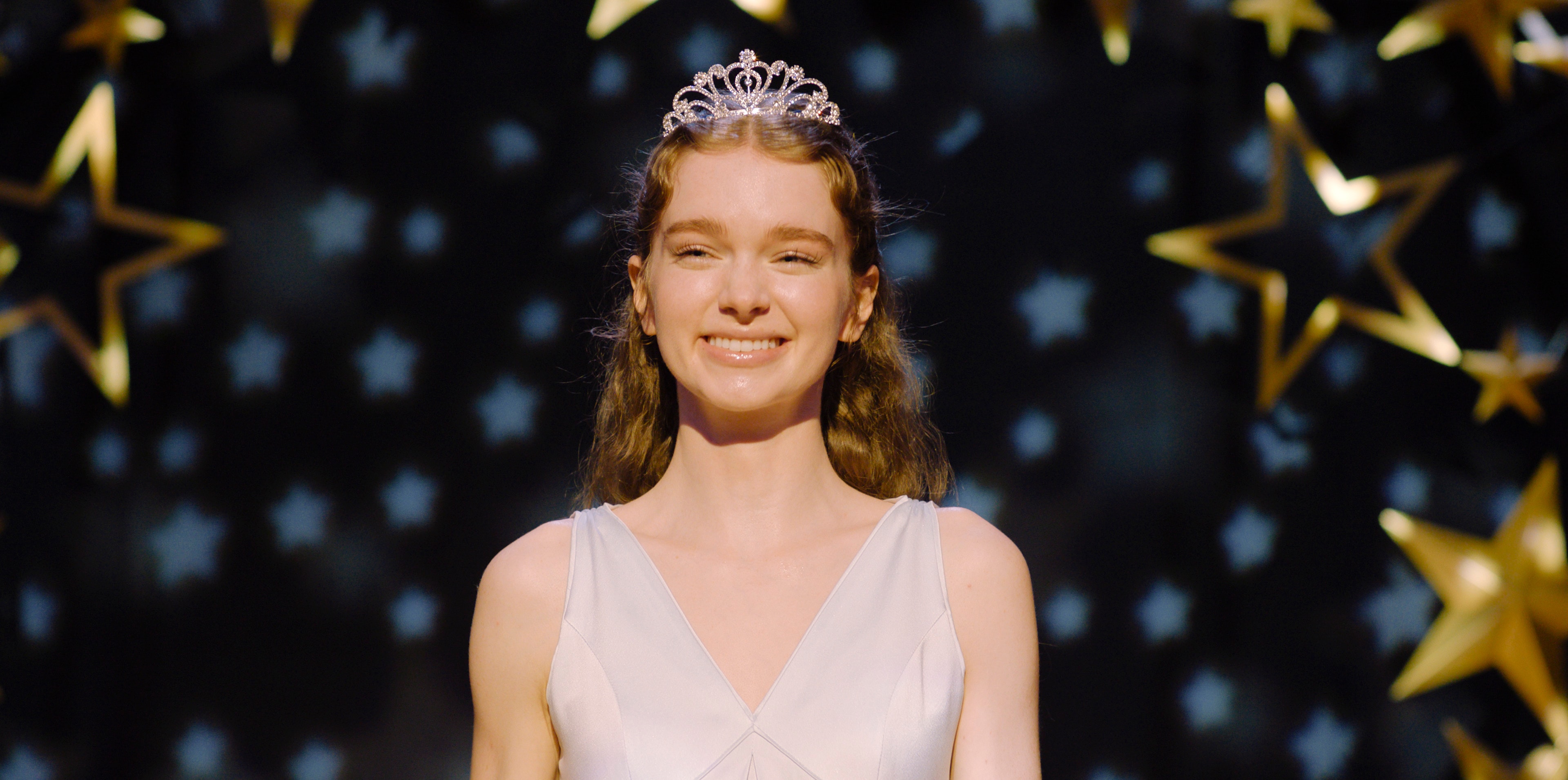 A smiling teenage girl wearing a silver tiara and a light-colored formal gown stands in front of a dark background decorated with gold stars.