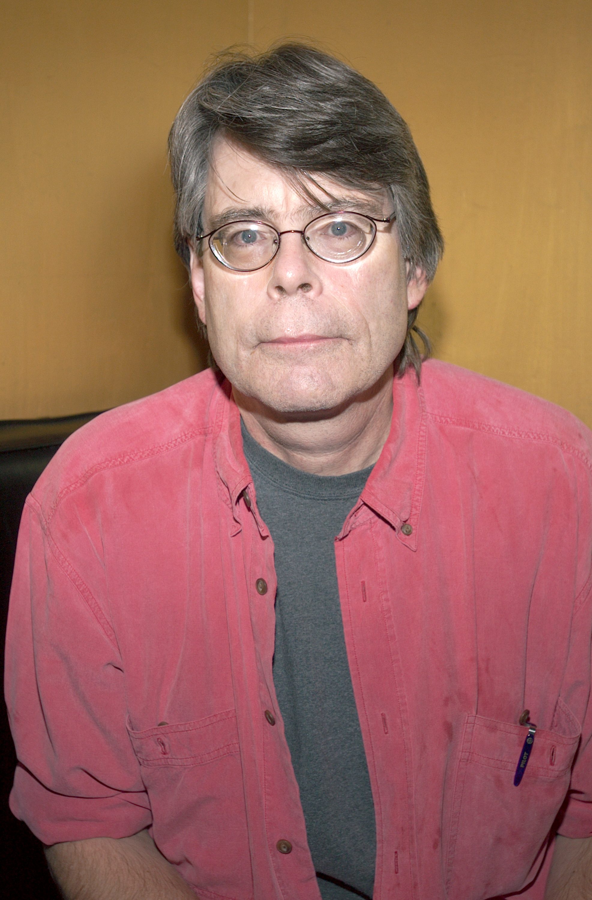 Stephen King, wearing round glasses and a red button-down shirt over a gray T-shirt, looks directly at the camera against a tan background.