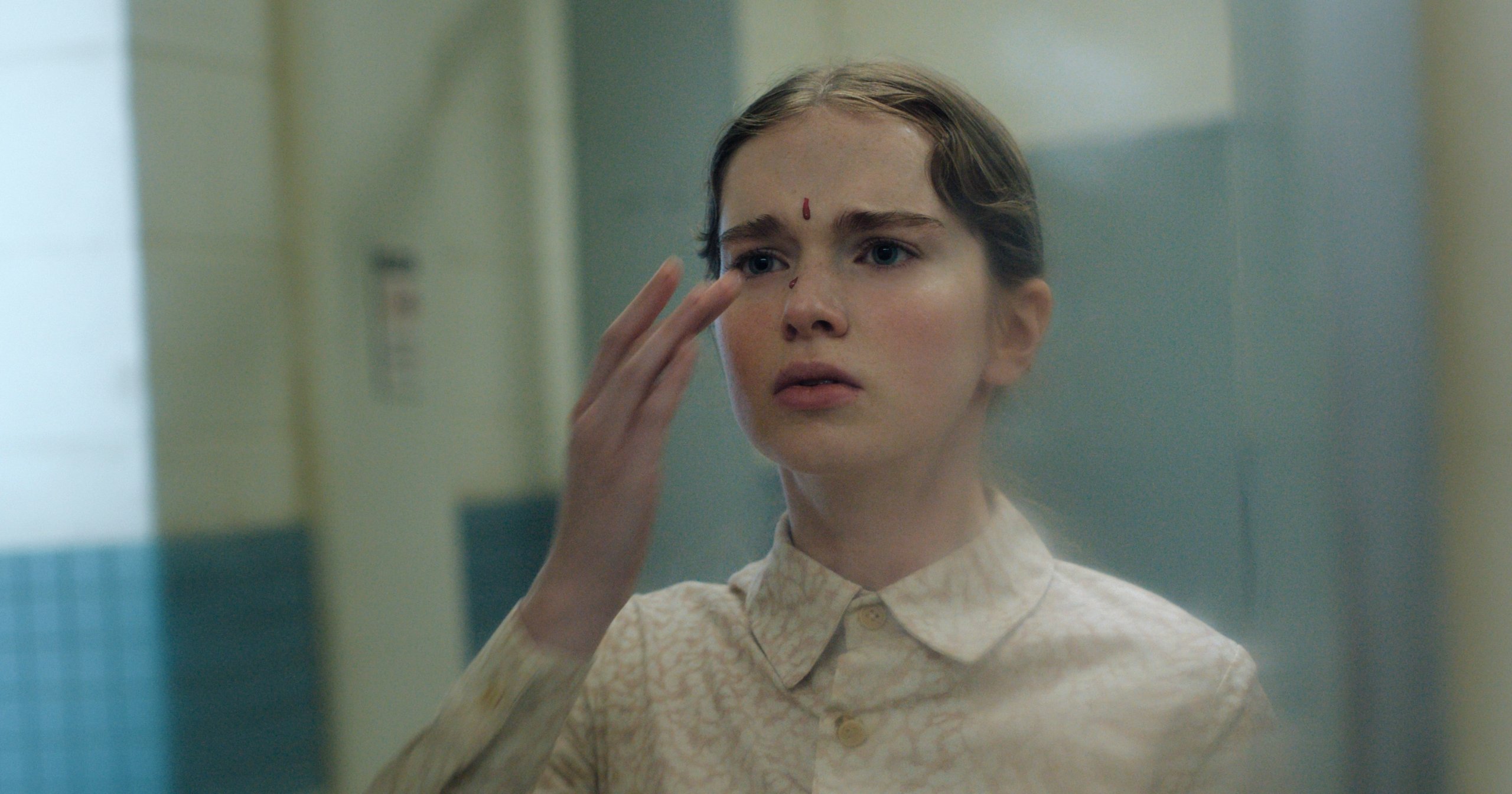 Young girl in a pale collared shirt with a drop of blood on her forehead, raising her hand to her face with a worried expression.