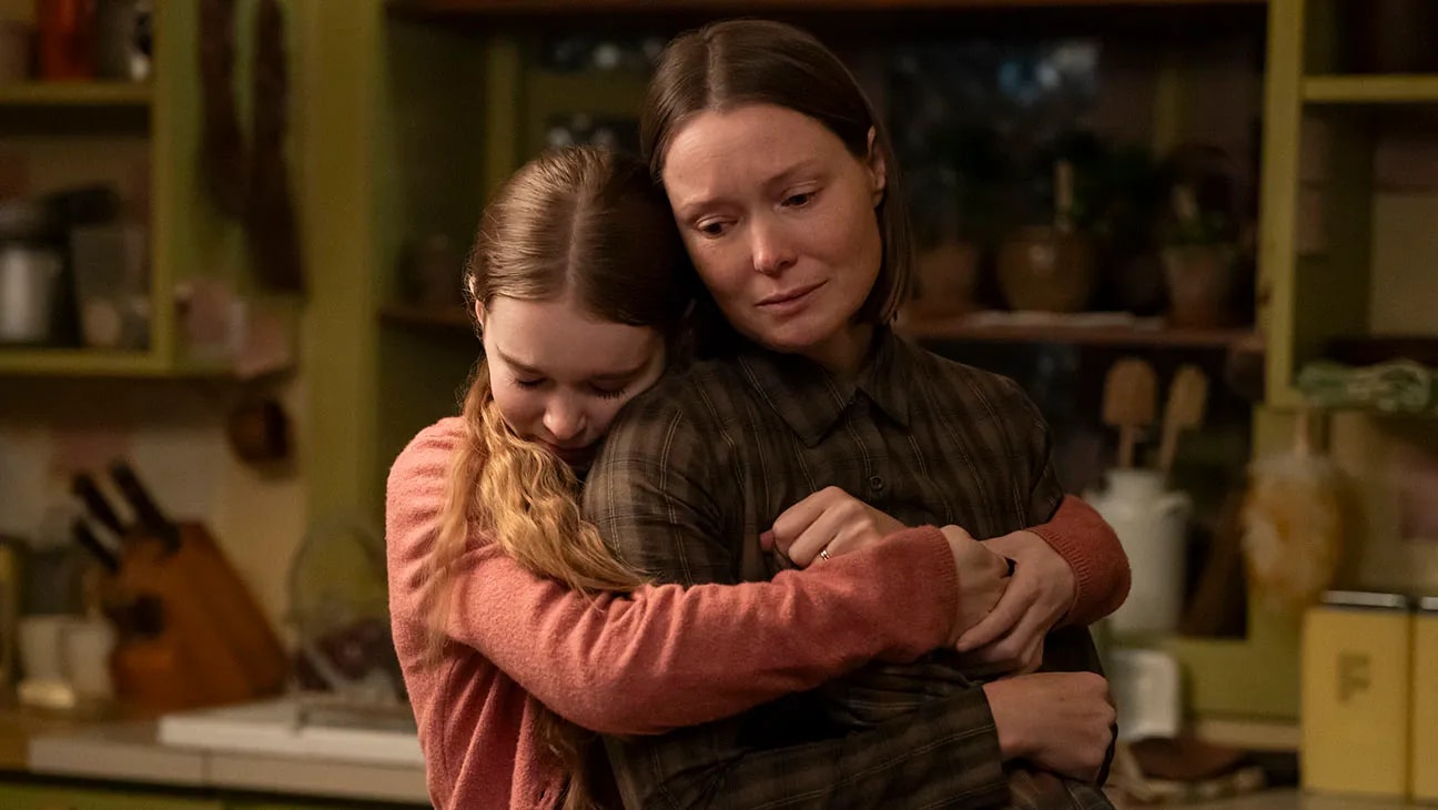 A teenage girl in a pink sweater hugs an older woman in a plaid shirt in a dimly lit kitchen, both with tearful expressions.