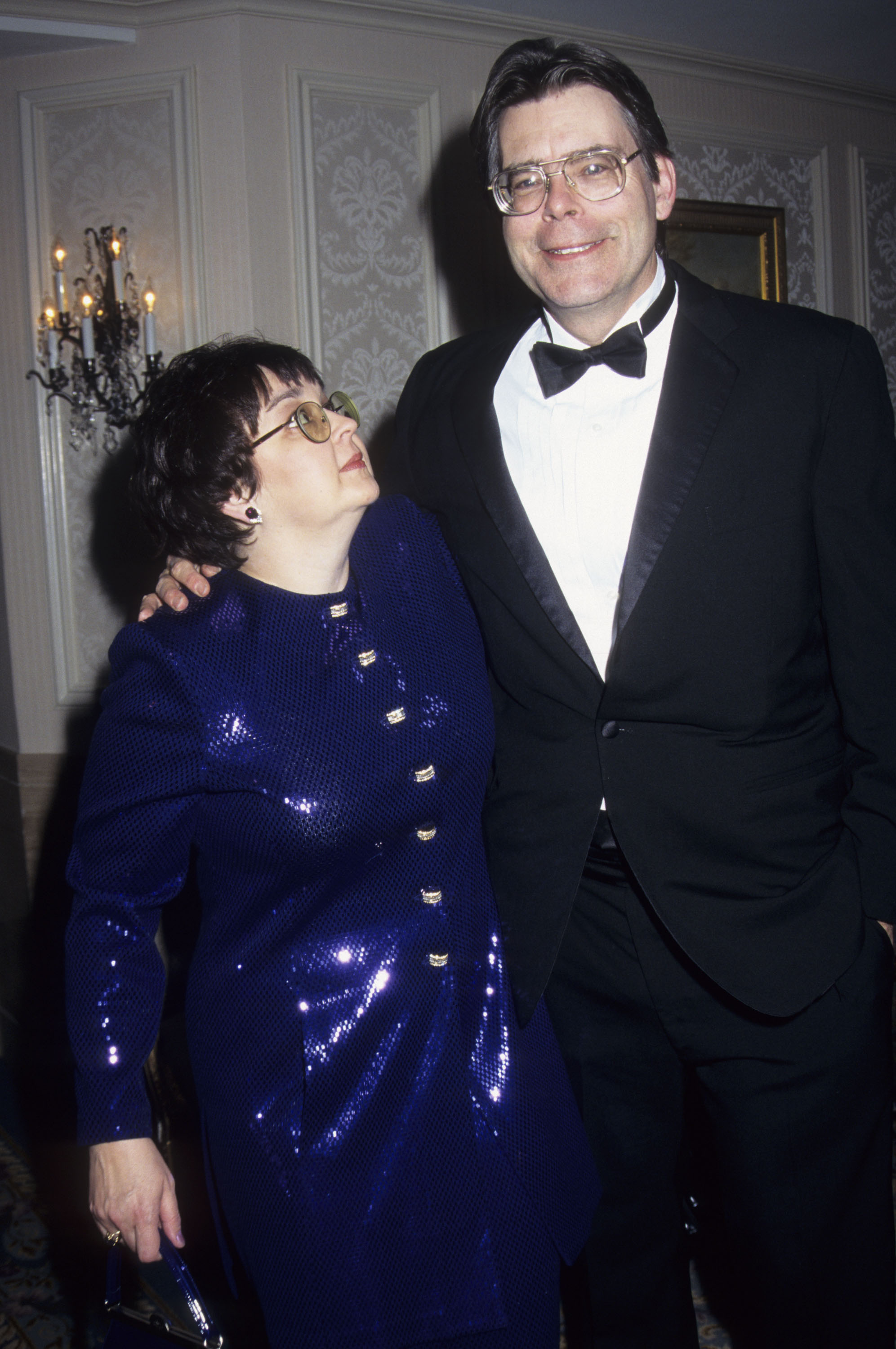 A tall man in a tuxedo with his arm around a shorter woman in a blue sequined dress, both wearing glasses, posing at a formal event.