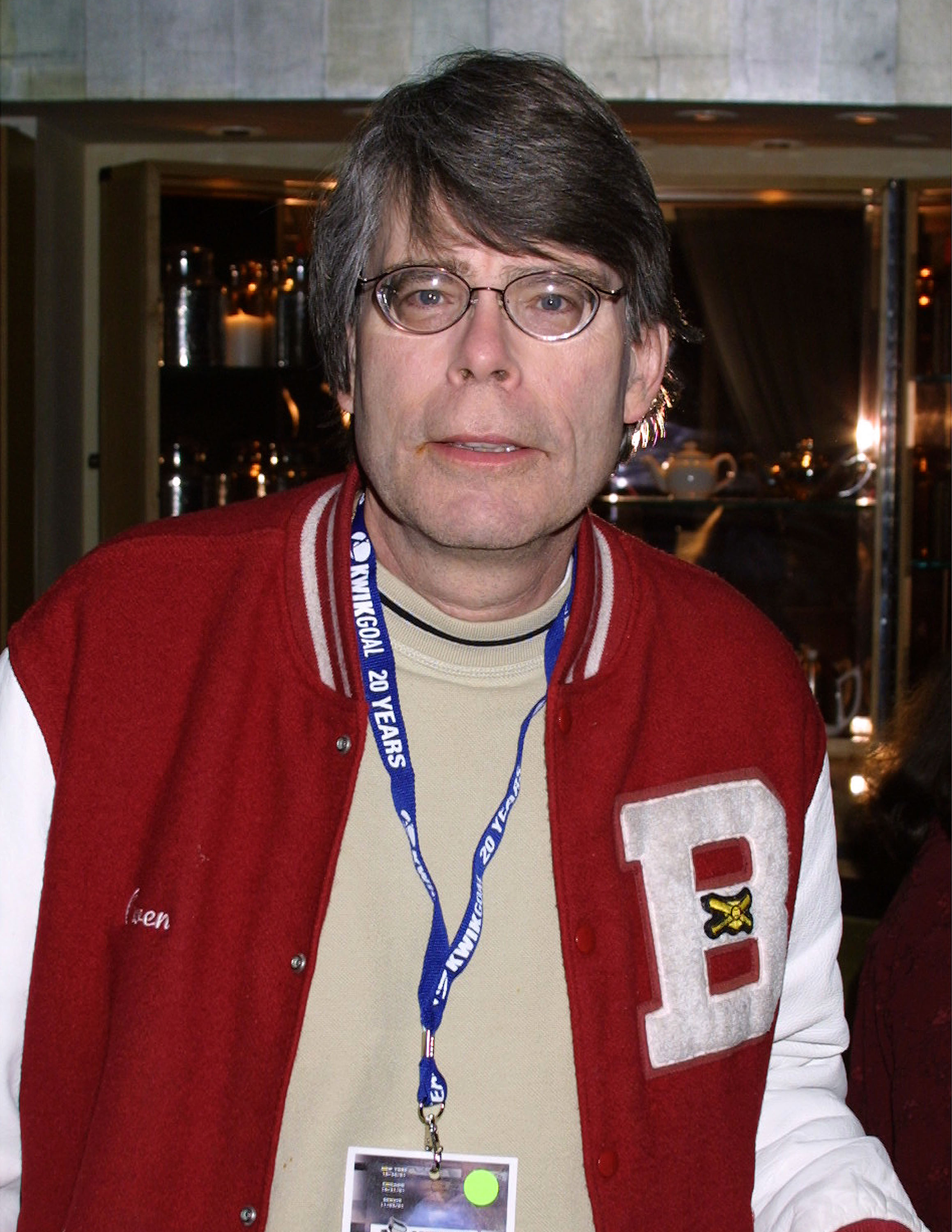 A man with dark hair and wire-rimmed glasses wearing a red varsity jacket with a 'B' letter and a blue lanyard at an indoor event.