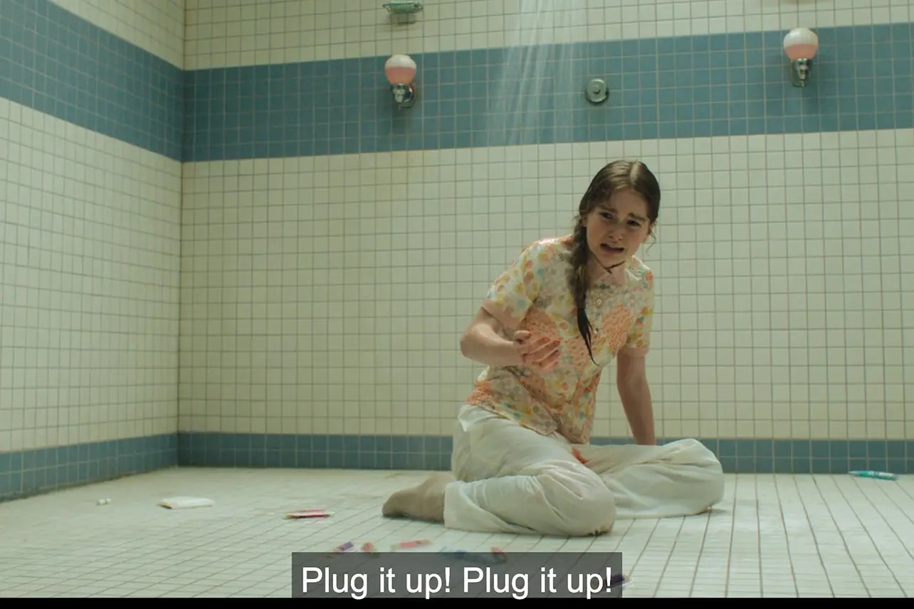 Young woman in a floral shirt sitting on a tiled shower room floor, hunched over and holding her stomach, with items scattered around her.