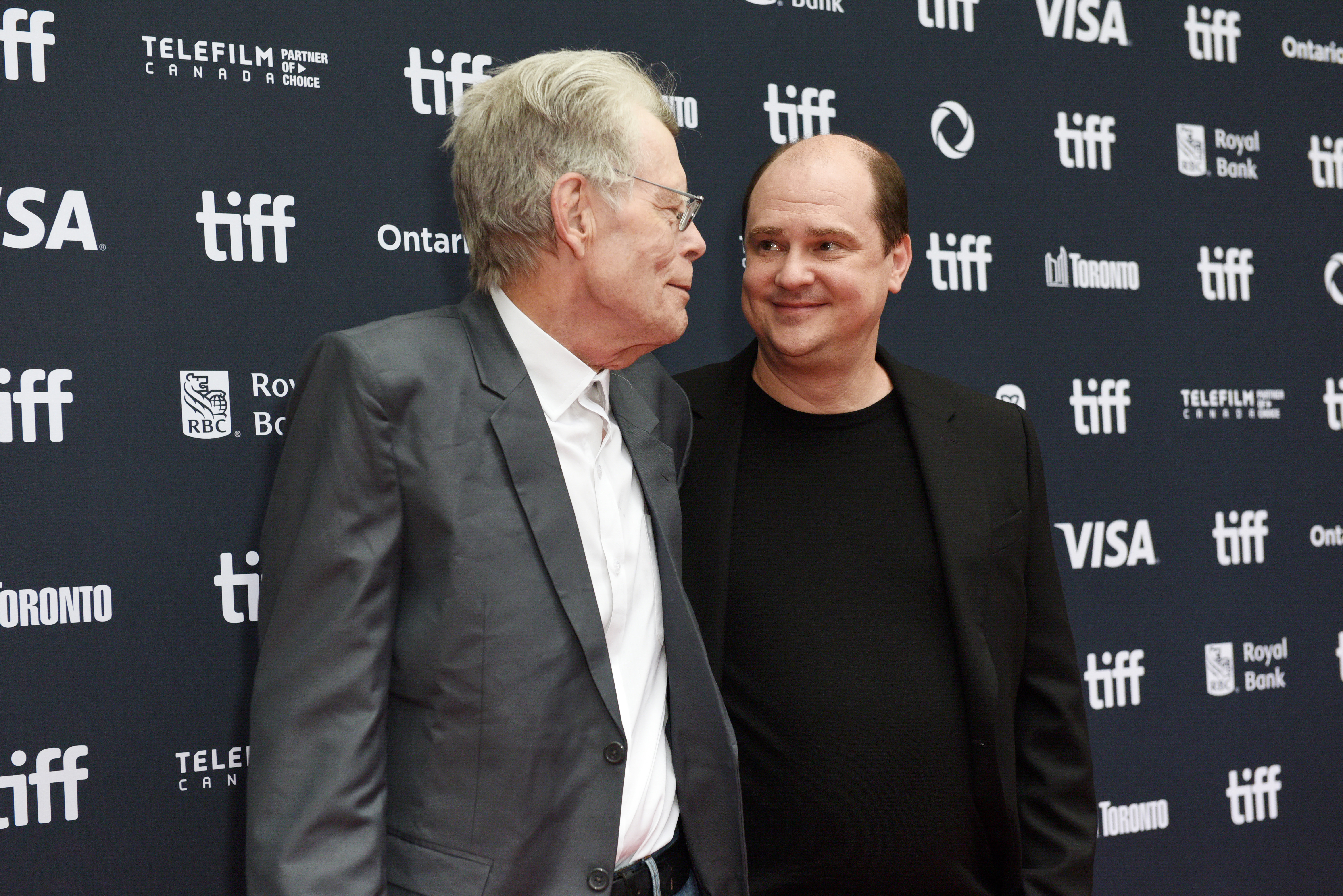 Stephen King and Mike Flanagan standing together at the TIFF 2024 red carpet backdrop, smiling and talking.
