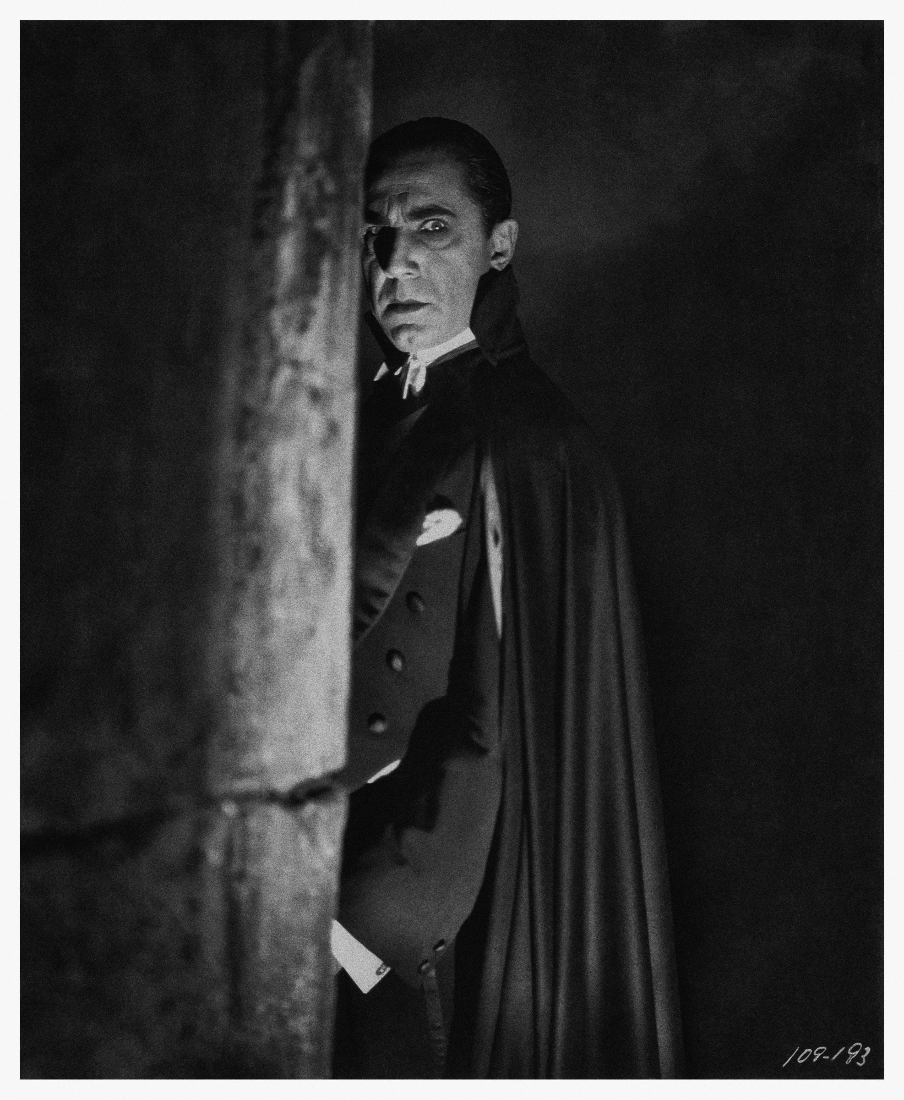 Black-and-white publicity still of Bela Lugosi as Dracula, wearing a cape and tuxedo, peering around a stone pillar in the 1931 film.