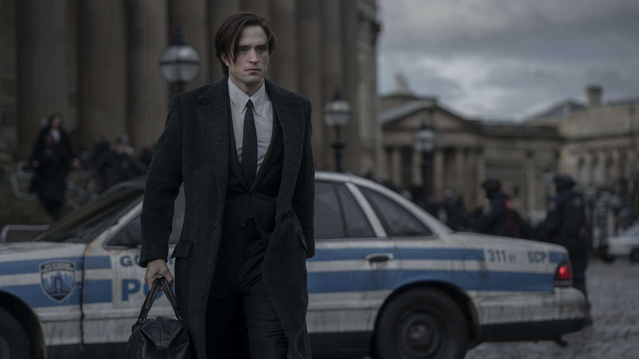A dark-haired man in a long dark coat and tie walks past white-and-blue police cars on a cobblestone street, with a grand building in the background.