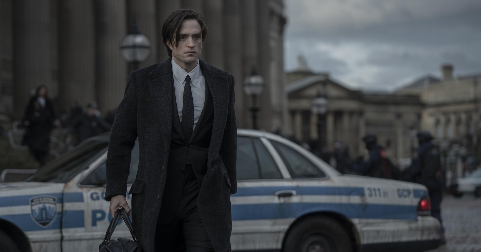 A dark-haired man in a long coat and tie carries a black bag past Gotham City police cars on a cloudy city street.