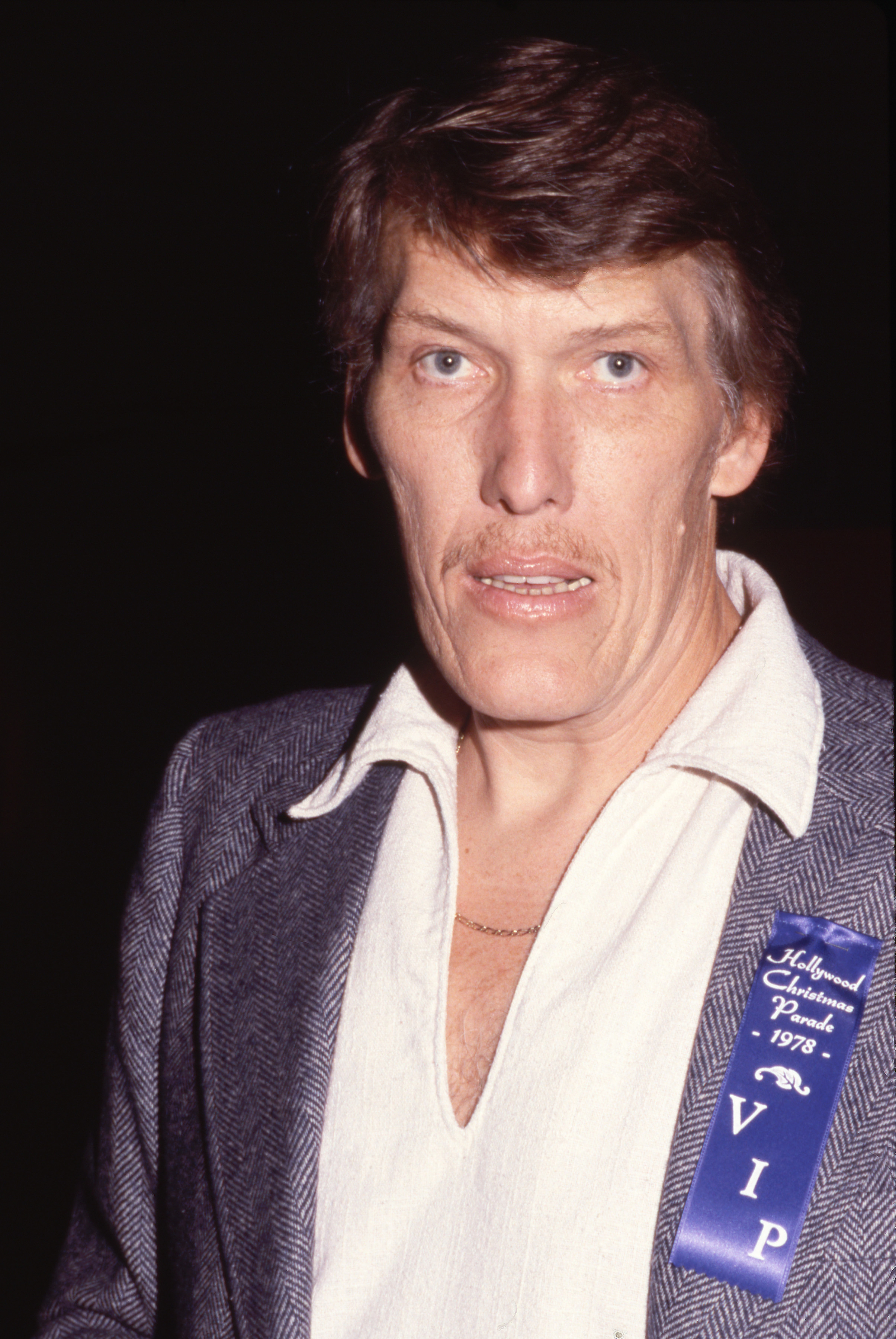Close-up of a man with dark hair and a mustache wearing a tweed jacket and a blue VIP ribbon reading 'Hollywood Christmas Parade 1978.'