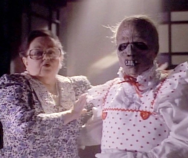 Click to open the image gallery A woman with glasses backs away from a horrifying zombie-like figure in a white polka-dot dress with red trim.