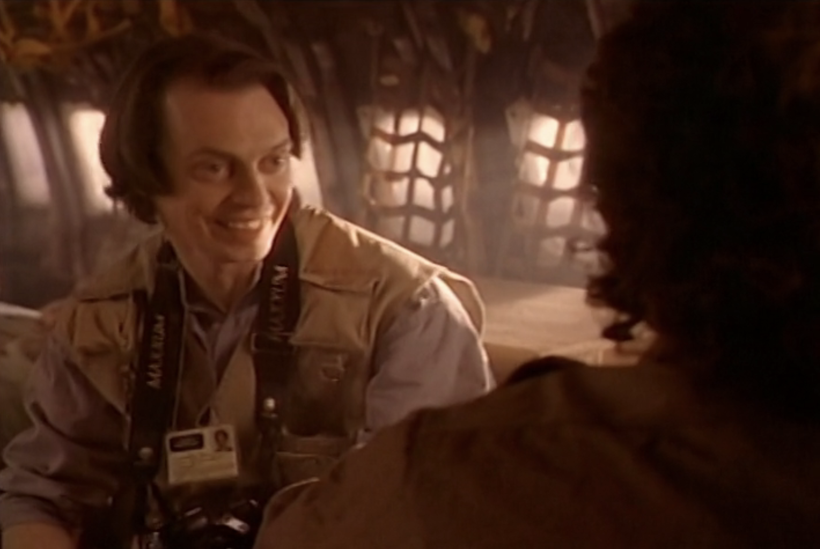 Click to open the image gallery Smiling man in a tan press vest with camera straps around his neck, seated inside a dimly lit military-style transport.
