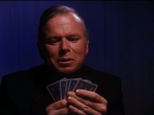 Click to open the image gallery Silver-haired man in black clothing holding and examining a fan of playing cards against a dark blue background.