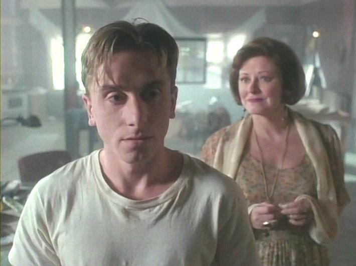 Click to open the image gallery Young man in a dirty white t-shirt in the foreground, older woman in a floral dress standing behind him in a hazy room.