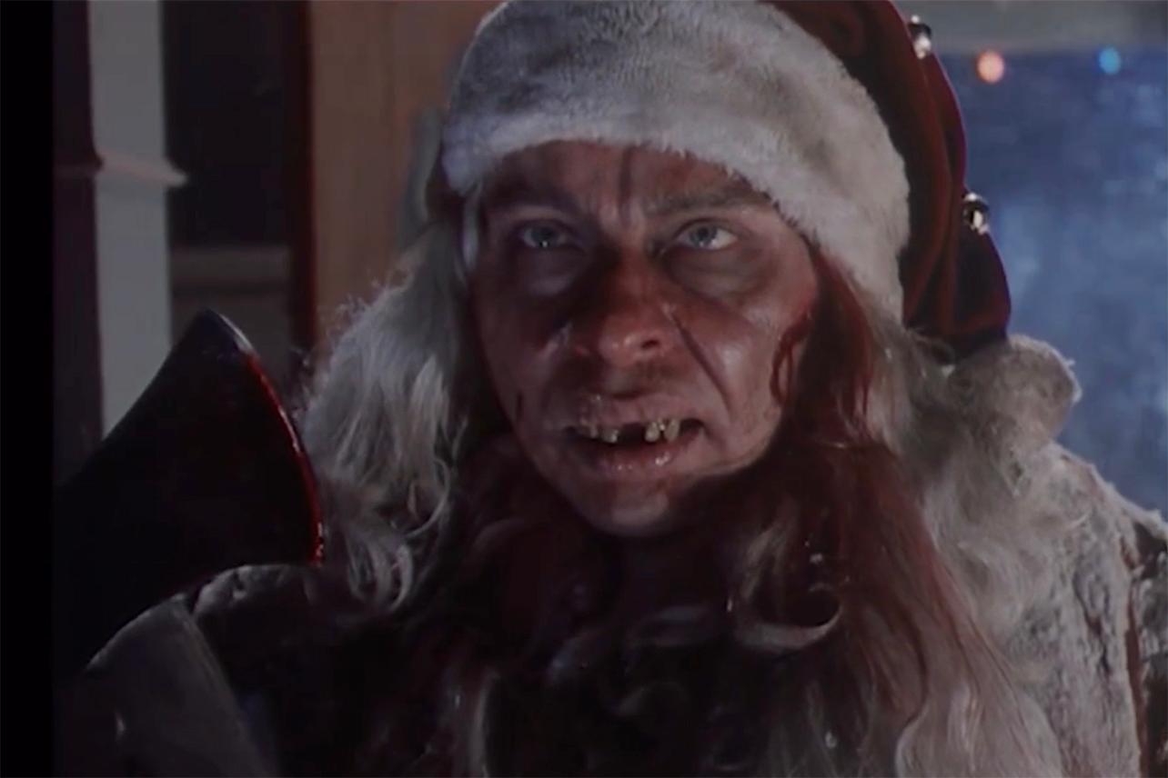 Click to open the image gallery A menacing, disfigured man with long gray hair, rotting teeth, and a Santa hat holds a bloody axe near a doorway at night.