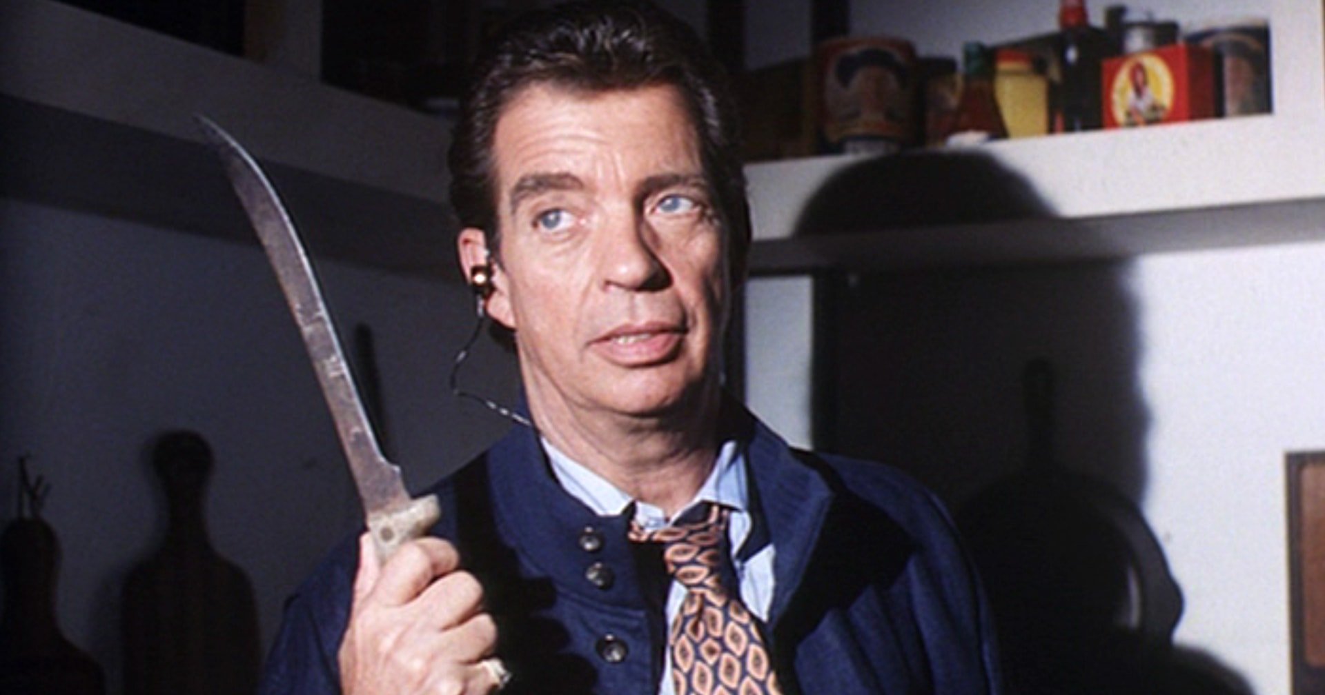 Man in a navy blazer and patterned tie holding a large knife, looking alarmed, in a dimly lit room with shelves in the background.
