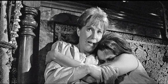 Black-and-white film still of two women huddled together in fear beside a bed post, one clutching the other protectively.
