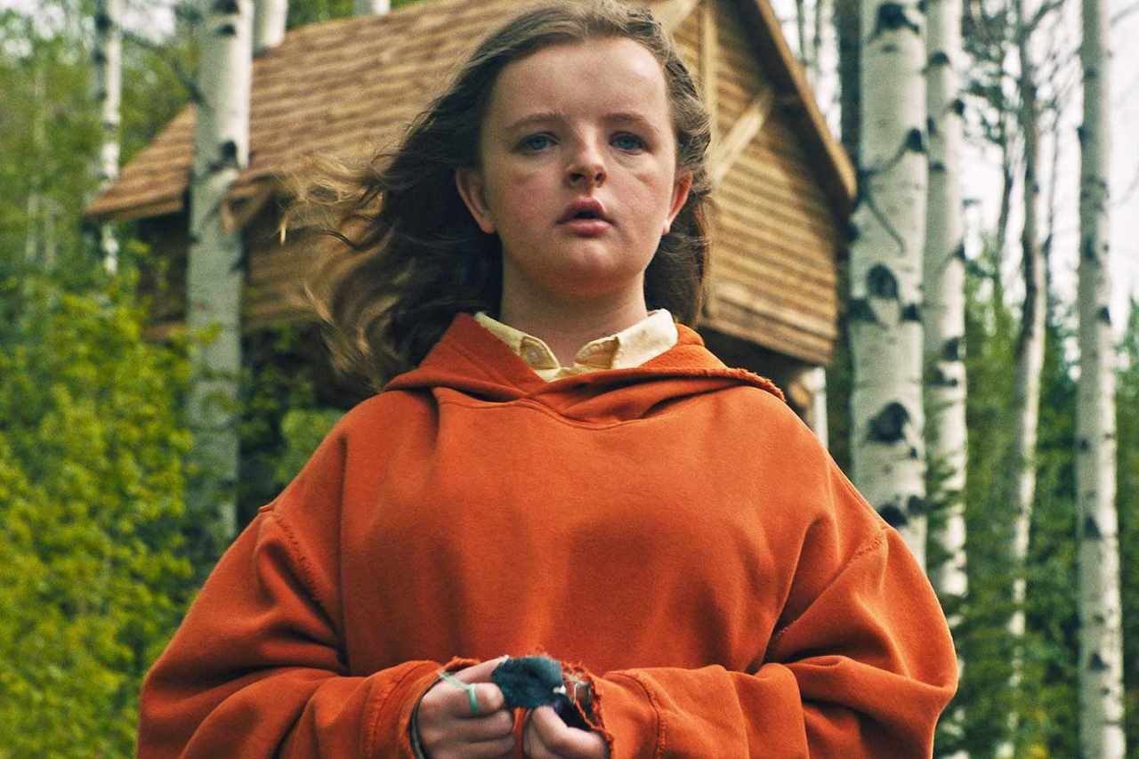 Young girl in an oversized orange hoodie stands among birch trees holding a small object, a wooden cabin visible behind her.