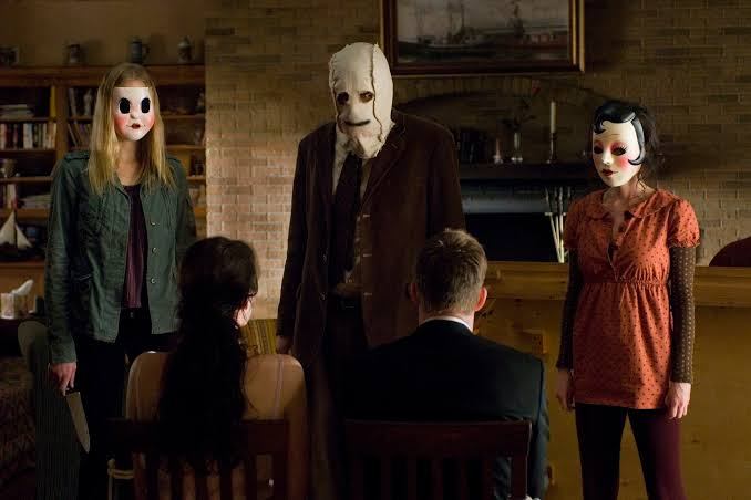 Three people wearing creepy masks stand over two seated people with their backs to the camera in a living room.