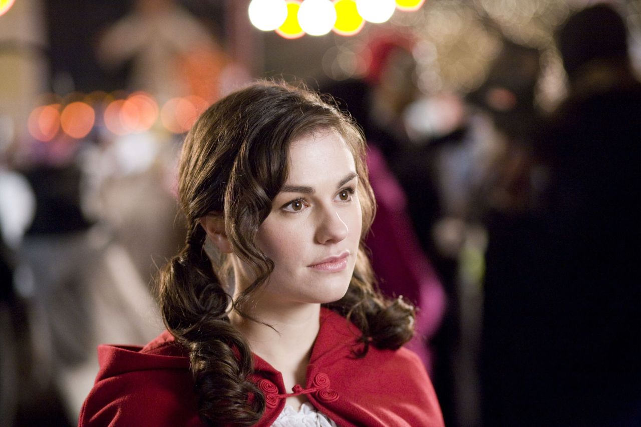 Young woman wearing a red hooded cape with curled brown hair looks off-camera, bokeh festival lights glowing behind her.