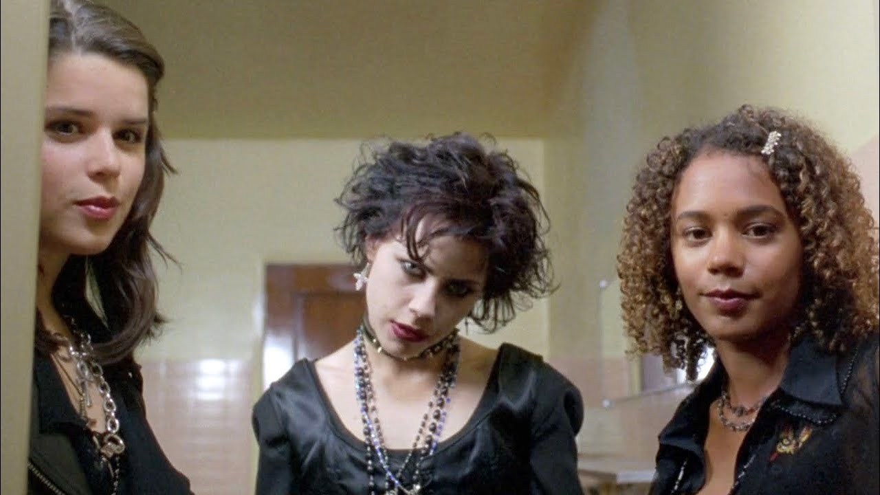Three teenage girls wearing black clothing stand close together in a school hallway, the middle girl wearing a choker and layered necklaces.