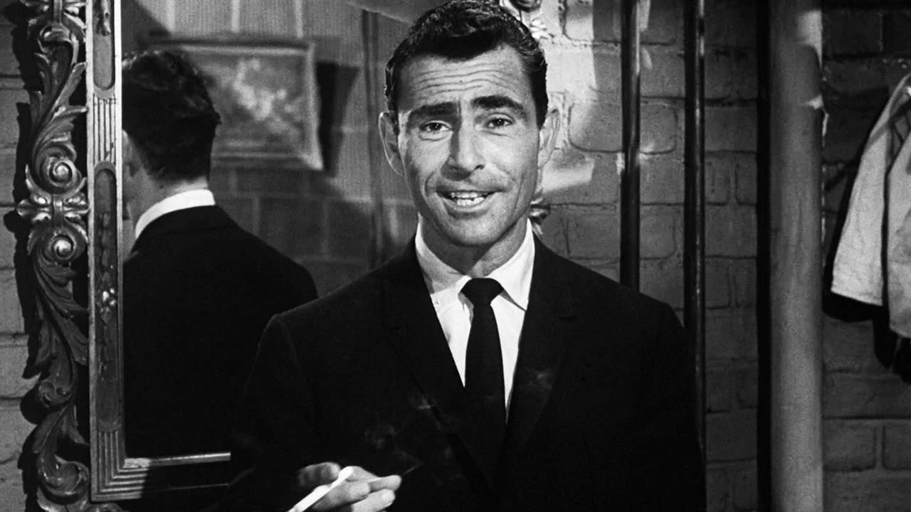Black-and-white image of a suited man holding a cigarette, speaking to camera beside an ornate gold-framed mirror.