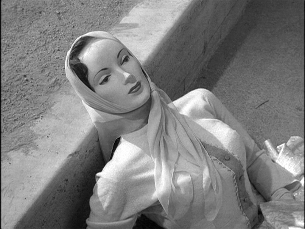 Black-and-white still of a woman in a head scarf looking upward, seated against a concrete wall.