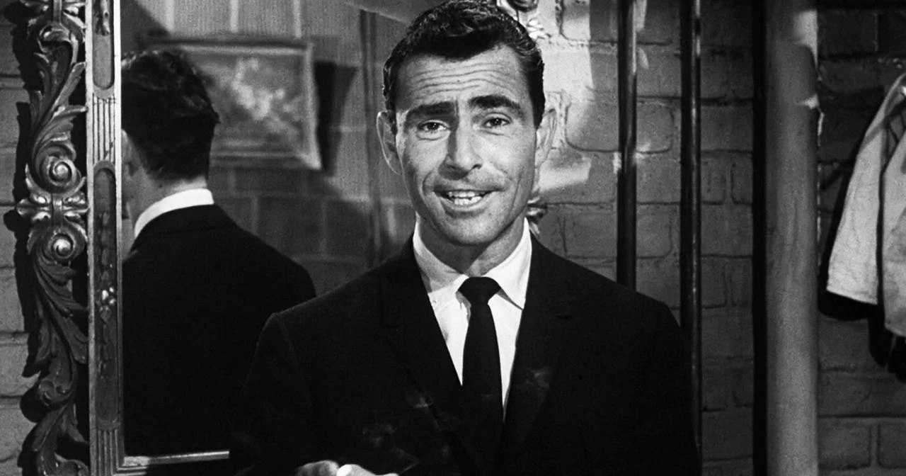 Black-and-white still of a man in a dark suit and tie holding a cigarette, standing in front of an ornate mirror.