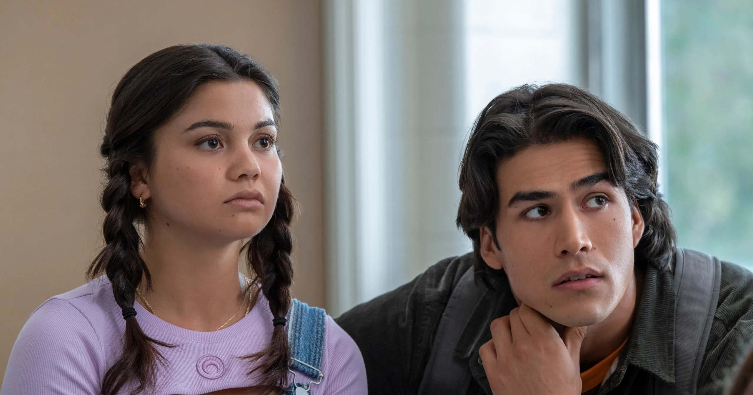 A young woman with dark braided hair in a lavender top and overalls sits beside a dark-haired young man in a jacket, both looking off to the side.