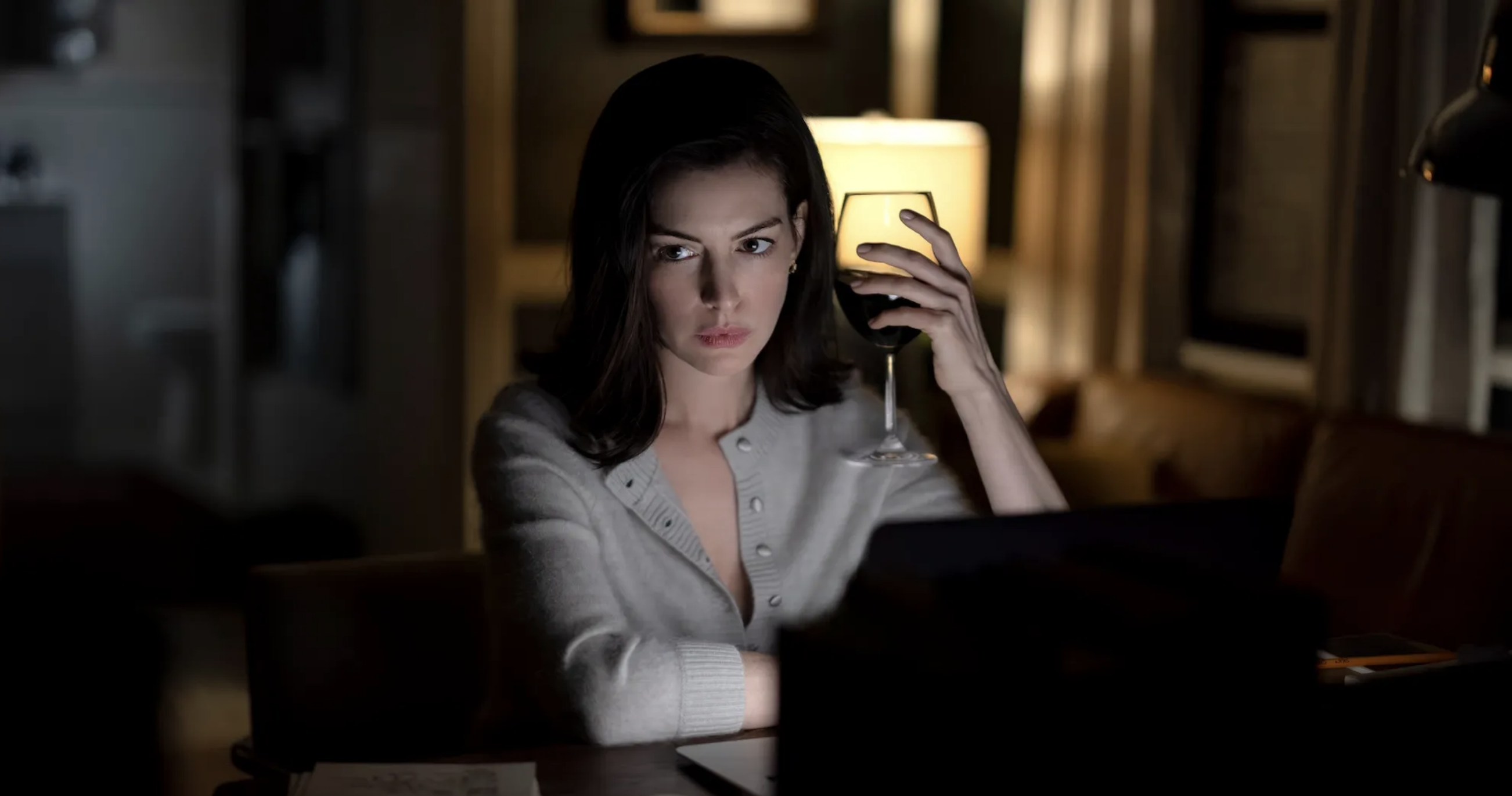Woman in gray cardigan holding a red wine glass, gazing at a laptop in a dark room lit by a warm lamp.