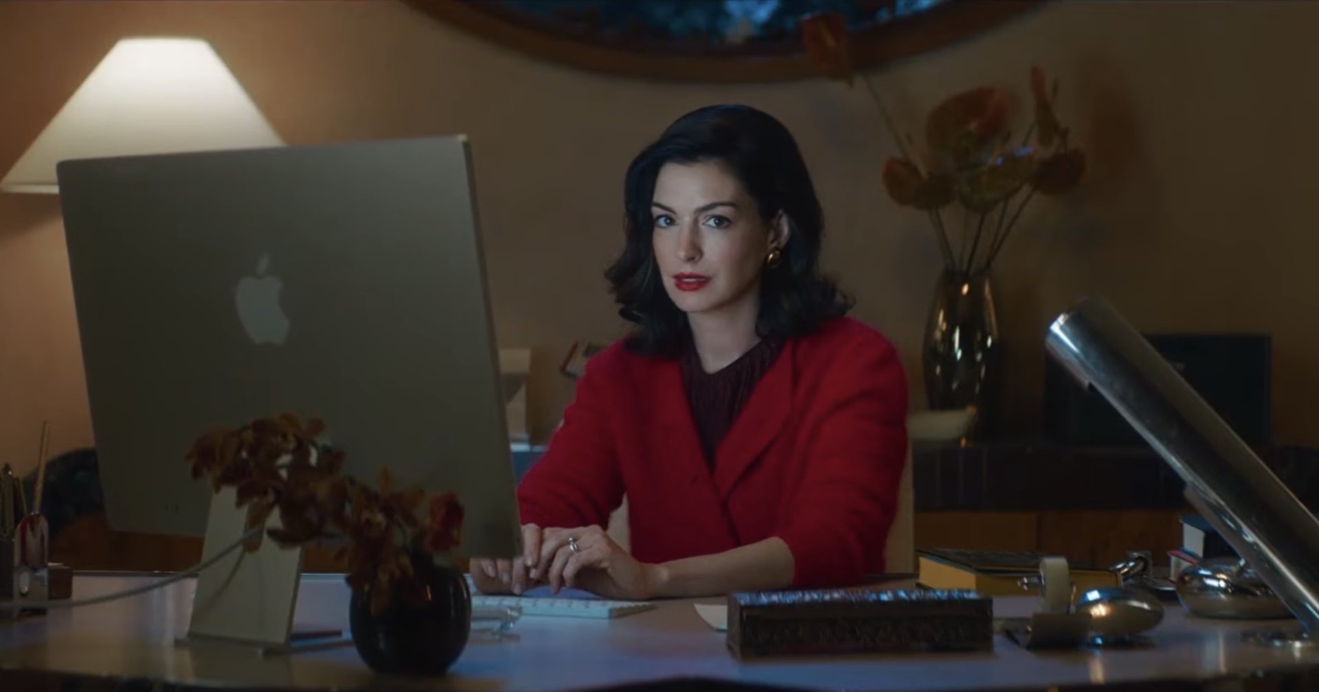 A dark-haired woman in a white cardigan holds a wine glass while gazing at a laptop screen in a dimly lit room.