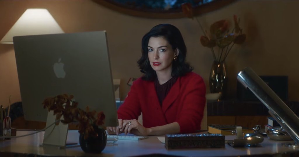 A dark-haired woman in a white cardigan holds a wine glass while gazing at a laptop screen in a dimly lit room.