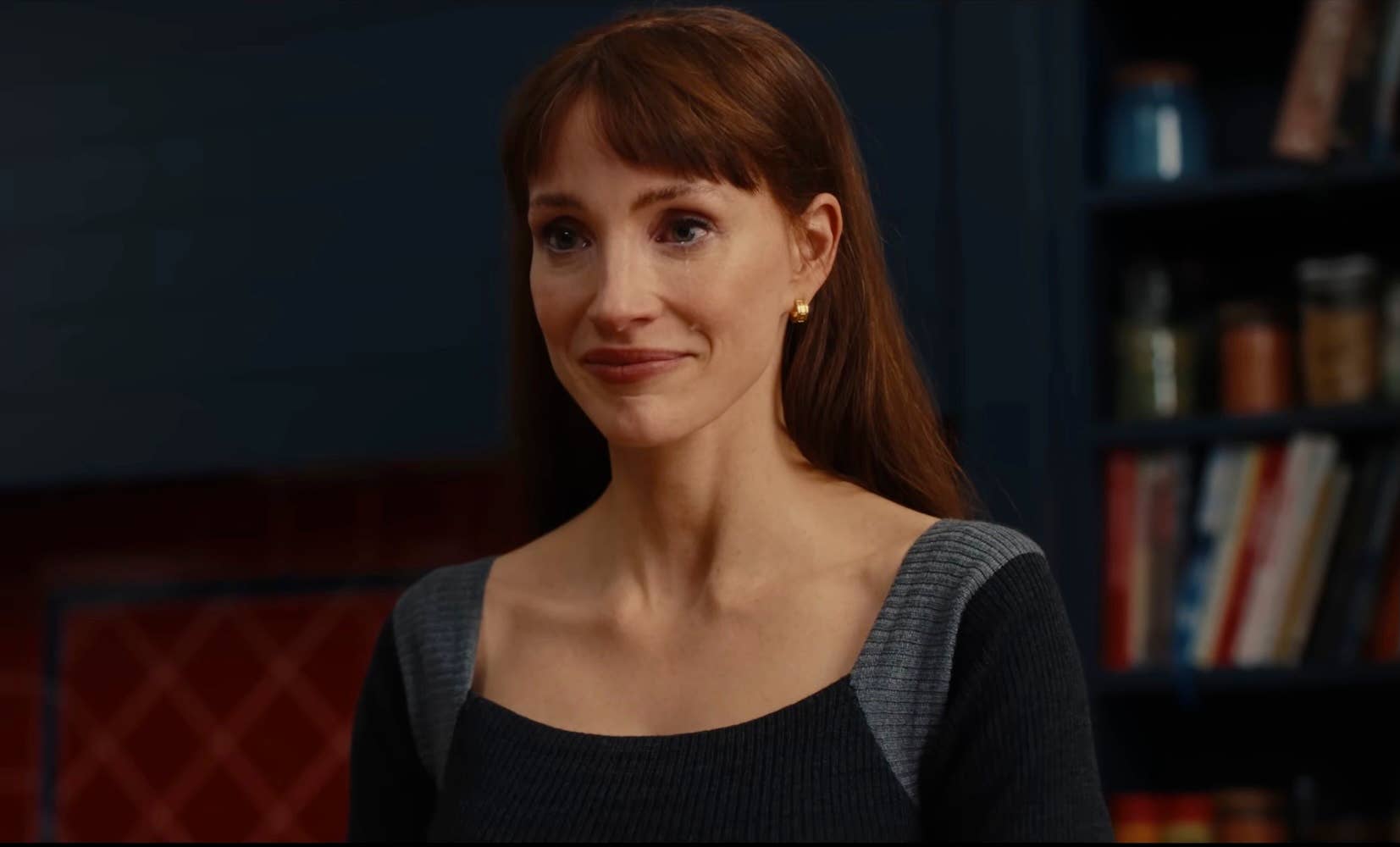 A red-haired woman with bangs wearing a gray ribbed top smiles slightly in front of a bookshelf in a dark-toned room.