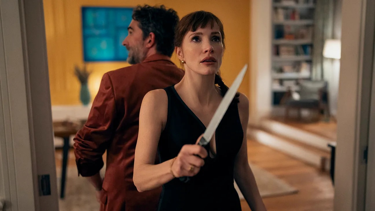 A red-haired woman in a black dress holds a large knife while standing back-to-back with a man in a rust-colored jacket in a warmly lit room.