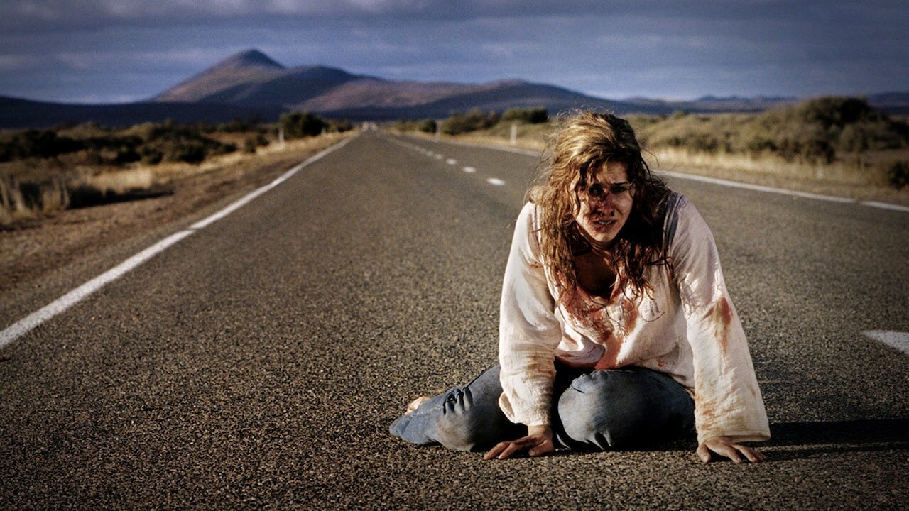 A bloodied young woman with tangled hair crawls on a remote two-lane road stretching toward distant mountains under a stormy sky.
