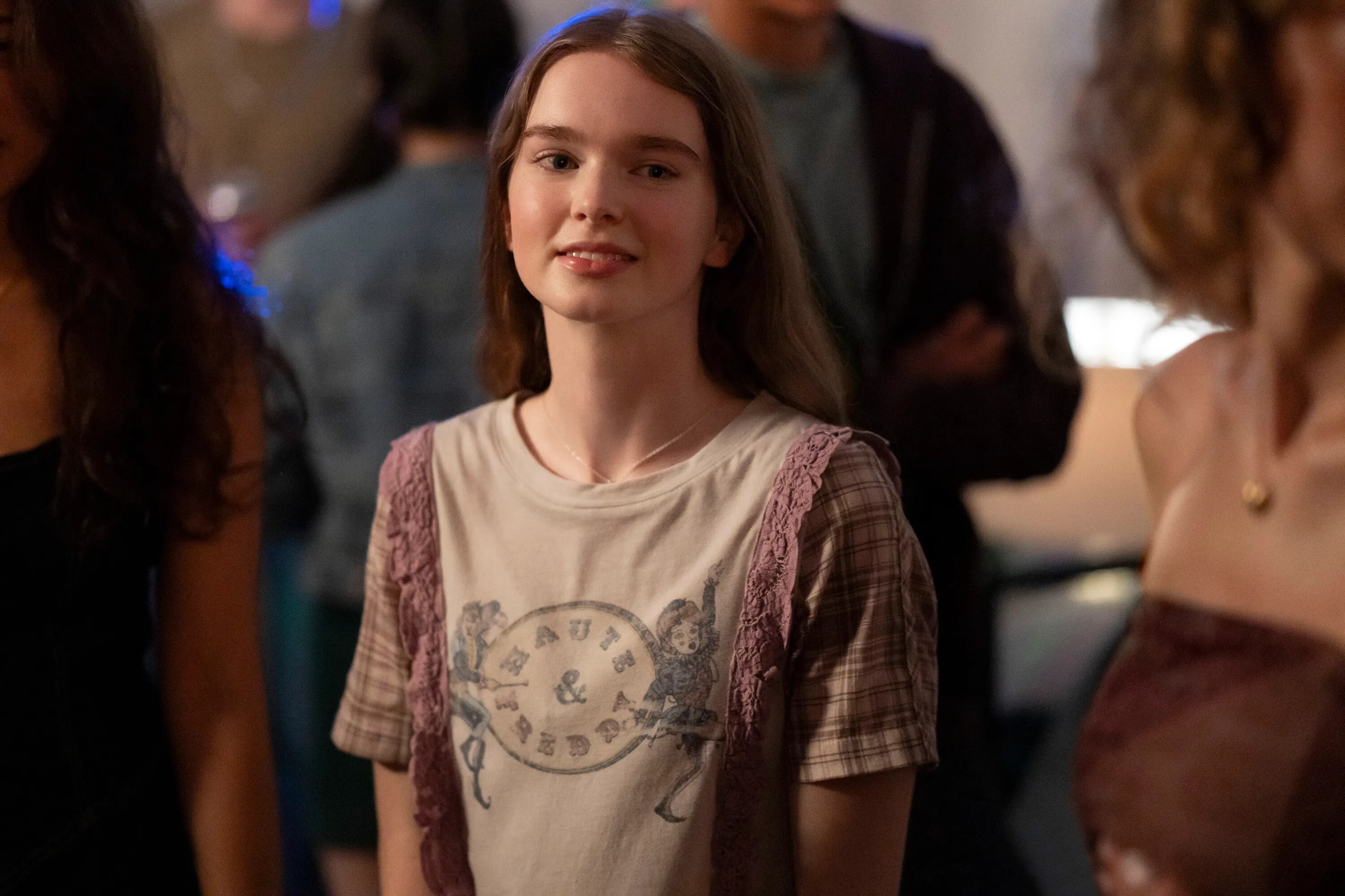 Young woman with long brown hair smiling softly in a graphic tee layered under a pink lace top, surrounded by blurred figures at a party.