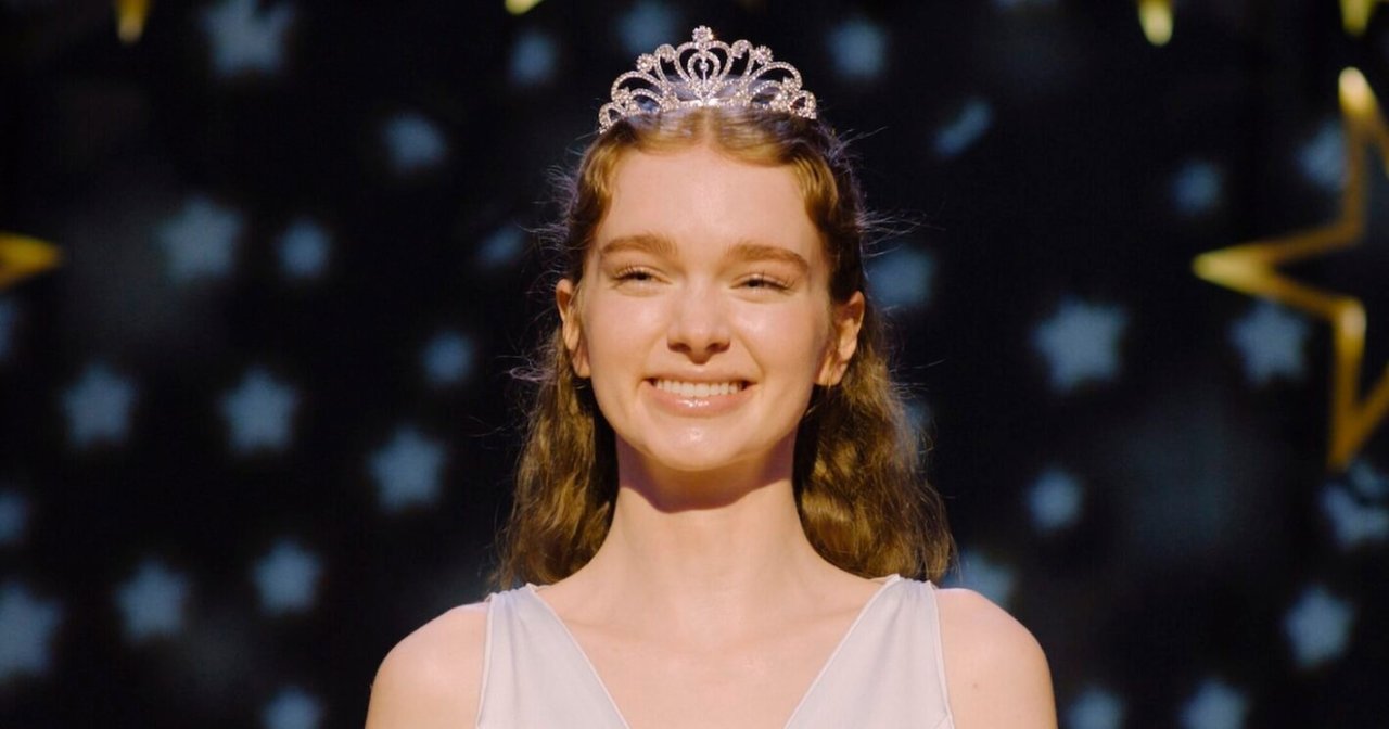 Young woman with auburn hair wearing a crystal tiara and white gown, smiling in front of a dark star-patterned backdrop.